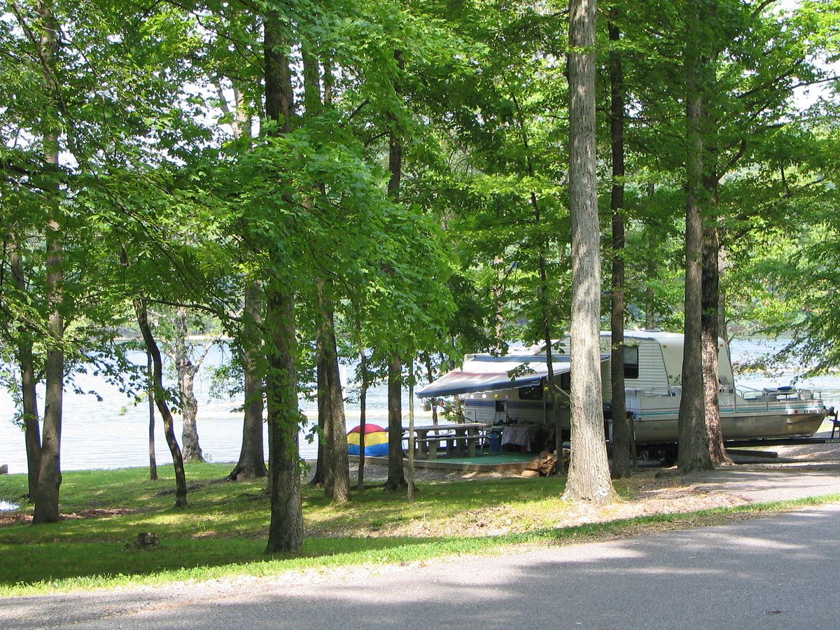 A travel trailer, picnic table and pontoon boat on a shaded lakefront site at Hillman Ferry Campground, Land Between the Lakes